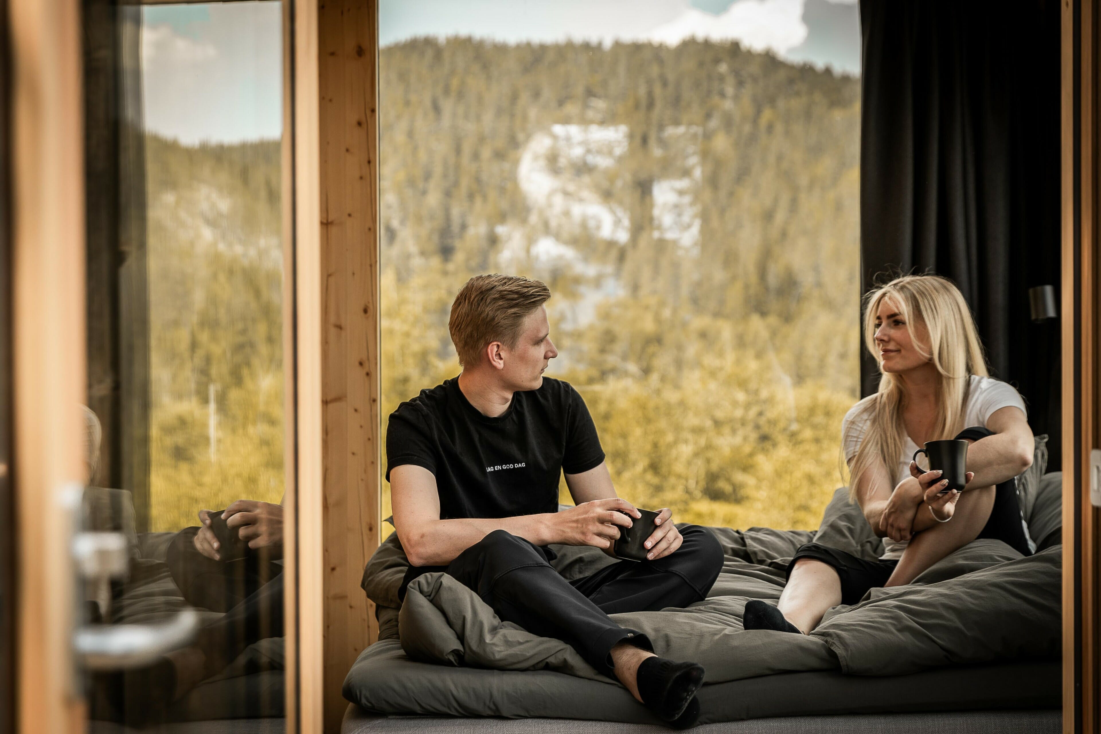 Et par sitte i senga og drikker kaffe, med utsikt fra panoramavinduer i bakgrunnen.